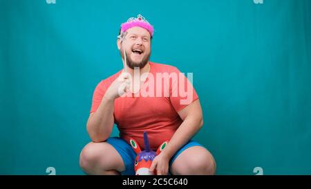 Un giovane uomo freaky con un braccio in una T-shirt rosa con un diadem sulla testa che cavalca un unicorno con una bacchetta magica in mano. Un divertente scherzo mago per fare e. Foto Stock