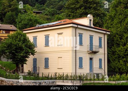 Bella vecchia casa ristrutturata con persiane blu, in legno, chiuse contro una fitta foresta verde nella regione Lombardia, Italia. Foto Stock