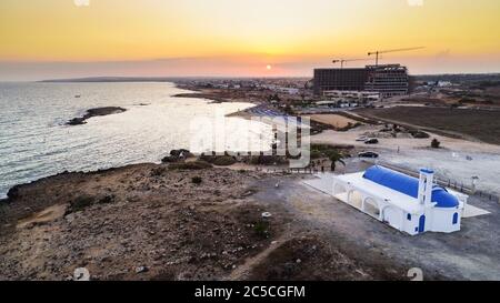 Vista aerea del tramonto sulla costa e tradizionale cappella dipinta di bianco con porte blu sulla spiaggia di Agia Thekla, Ayia Napa, Famagosta, Cipro da A. Foto Stock