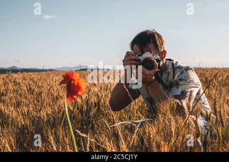 Bel giovane scatta una foto con la sua fotocamera di un fiore. È accovacciato nel mezzo di un campo di grano. Foto Stock