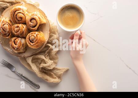 vista dal taglio corto della donna che tiene una tazza di caffè vicino a rotoli di cannella fatti in casa sulla superficie in marmo con forchetta e panno Foto Stock