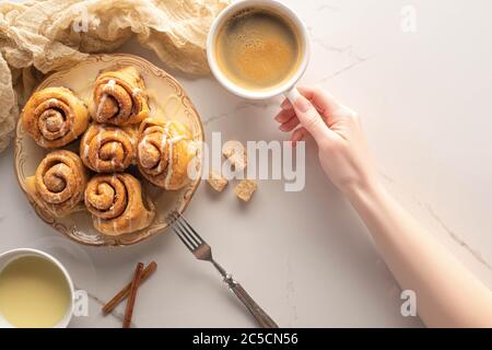 vista dal taglio corto della donna che tiene una tazza di caffè vicino a rotoli di cannella fatti in casa sulla superficie di marmo con forchetta, latte condensato e panno Foto Stock