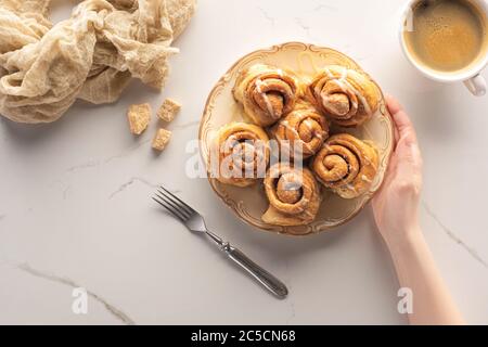 vista dal taglio corto di una donna che tiene rotoli di cannella fatti in casa sulla superficie in marmo con forchetta, tazza di caffè e panno Foto Stock