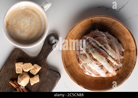 vista dall'alto del fresco rotolo di cannella fatto in casa su un piatto sulla superficie di marmo con tagliere con zucchero di canna, bastoncini di cannella vicino al caffè Foto Stock