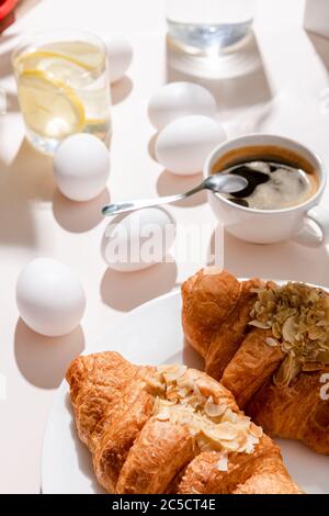 Uova sode, due croissant, tazza di caffè e un bicchiere d'acqua con limone per la colazione su un tavolo grigio con ombre Foto Stock