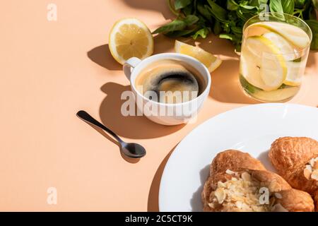 caffè, croissant e bicchiere d'acqua con limone e basilico per la colazione su un tavolo beige Foto Stock