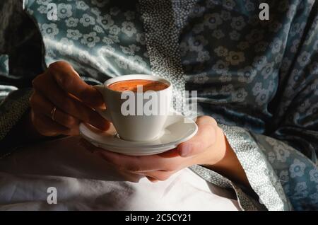 Caffè mattutino a letto. Una tazza di caffè in mani femminili al sole del mattino. Godendo le piccole cose. Primo piano Foto Stock