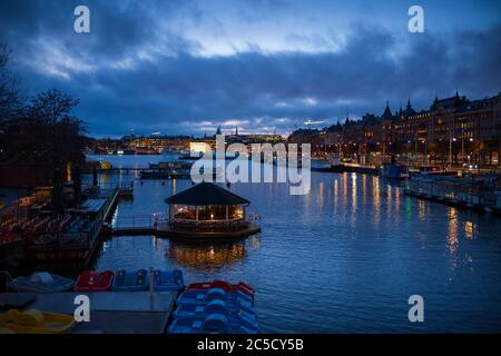 Durante un tramonto, le luci della città di Stoccolma, Svezia, brillano, viste dal ponte Djurgardsbron che si affaccia su un canale pieno di barche ormeggiate. Foto Stock