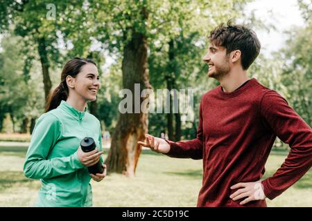 Un sportivo sorridente che parla con la ragazza con una bottiglia sportiva nel parco Foto Stock