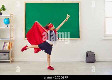 Studentessa in un costume da supereroe che si pone sullo sfondo di una scuola verde in una classe scolastica. Foto Stock