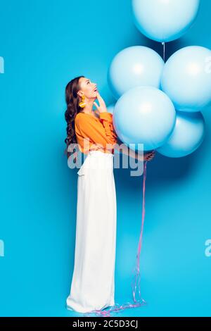 donna elegante e sbalordita che tocca il mento mentre si guarda su grandi palloncini festive su sfondo blu Foto Stock