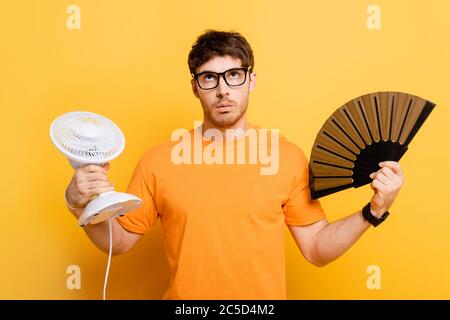 dispiaciuto giovane uomo che tiene ventilatori elettrici e mani mentre si guarda in su sul giallo Foto Stock