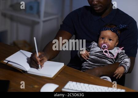 Padre africano che prende appunti in blocco note mentre si siede al tavolo con il bambino sulle ginocchia Foto Stock