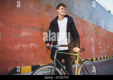 Giovane uomo con capelli marroni che cammina con la classica bicicletta mentre si guarda da parte con gioia. Ragazzo sorridente in giacca e zaino nero con Foto Stock