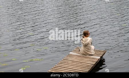 Giovane donna seduta sul molo di legno, riposante e guardando il fiume, vista dall'alto. Ora legale. La gioia della vita di campagna. Foto Stock