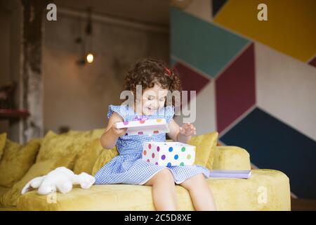 Gioiosa bambina con capelli ricci scuri in abito felicemente apertura confezione regalo sul divano a casa Foto Stock