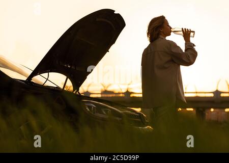 Silhouette di donna che beve acqua da una bottiglia ricaricabile, stanco di aspettare l'assistenza vicino alla sua auto rotto con cofano aperto al tramonto. Foto Stock