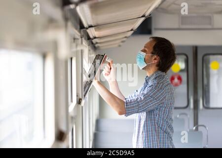 Uomo in camicia controllata apre una finestra su un treno per respirare aria fresca e ventilazione, indossando maschera medica viso durante il nuovo cambiamento normale dopo corona Foto Stock