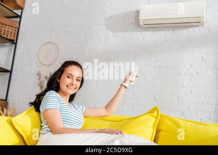 Giovane donna sorridente alla telecamera mentre si accende il condizionatore d'aria con il telecomando sul divano Foto Stock