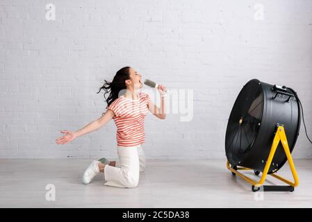 Vista laterale della giovane donna che canta e tiene la spazzola mentre si inginocchiava vicino alla ventola elettrica a casa Foto Stock