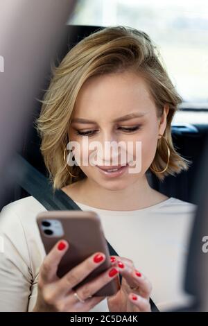 Donna sorridente seduto in auto sul sedile passeggero allacciato da una cintura di sicurezza, utilizzando smartphone, scorrimento dei social network, lettura dei messaggi. Primo piano. Foto Stock