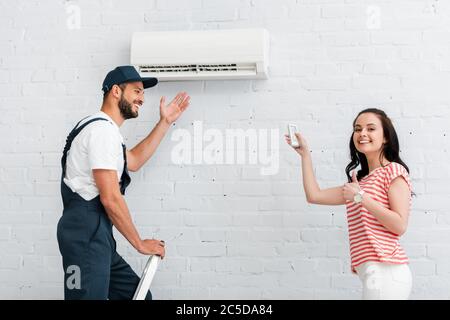 Vista laterale di una donna sorridente con telecomando del condizionatore d'aria che mostra il pollice in alto vicino al workman in tute sulla scala Foto Stock