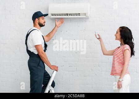 Vista laterale del workman che punta al condizionatore d'aria vicino a una donna sorridente che tiene il telecomando Foto Stock