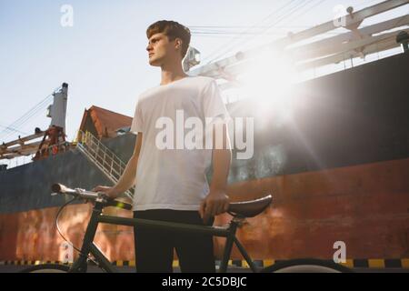 Ragazzo serio con i capelli marroni in piedi con la bicicletta e guardando da parte con attenzione. Giovane uomo in maglietta bianca in piedi e in posa sulla macchina fotografica con Big Foto Stock