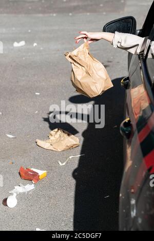 Donna che tiene in mano il sacchetto di carta dei rifiuti, fuori dal finestrino che guida in auto. Autista che getta rifiuti nel parcheggio. Inquinamento ambientale Foto Stock