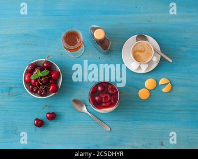 Panna cotta dessert tradizionale italiano con ciliegie in camicia al rum e sciroppo di miele. Tavolo di legno blu, ciotola delle ciliegie fresche, bicchiere di panna co Foto Stock