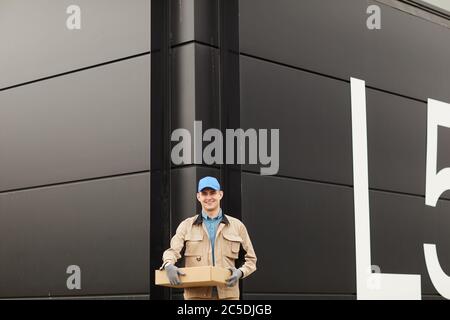 Ritratto di giovane corriere in uniforme tenendo pacco e sorridendo alla fotocamera mentre si sta in piedi contro il magazzino all'aperto Foto Stock