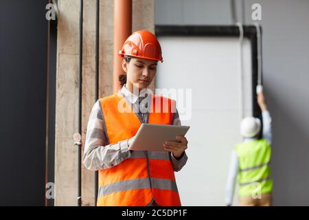Giovane donna in casco da lavoro e in gilet riflettente con tablet digitale mentre lavora nel cantiere Foto Stock