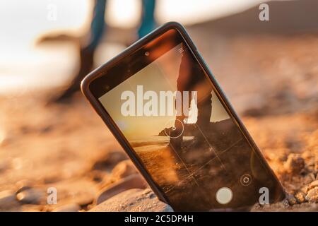 Il concetto delle foto sul telefono. Lo smartphone è sepolto nella sabbia sulla spiaggia e scatta una foto di un uomo sullo sfondo. Primo piano. Foto Stock