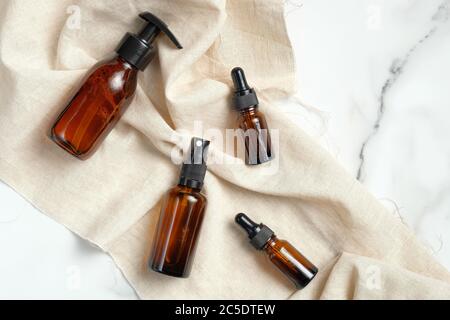 Set di bottiglie cosmetiche in vetro ambrato con tessuto beige su tavolo in marmo. Disposizione piatta, vista dall'alto. Prodotti di bellezza per la cura della pelle dell'uomo, cosmetici naturali organici Foto Stock