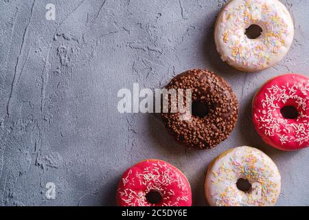 Ciambelle di cioccolato, rosa e vaniglia con spruzzi, dessert con cibi dolci smaltati su fondo testurizzato in cemento, spazio per la copia vista dall'alto Foto Stock