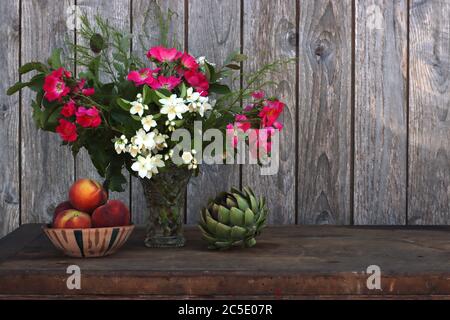Un vaso di fiori estivi accanto a una ciotola in ceramica fatta a mano piena di pesche e carciofo Foto Stock