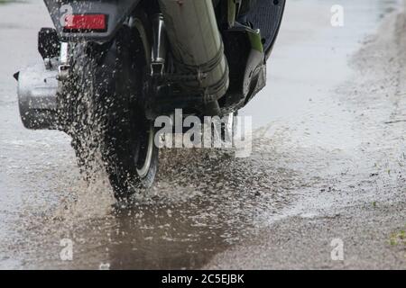 Moto ciclomotore si muove attraverso un puddle su una strada bagnata sotto la pioggia. Lo spruzzo vola dalle ruote. Primo piano Foto Stock