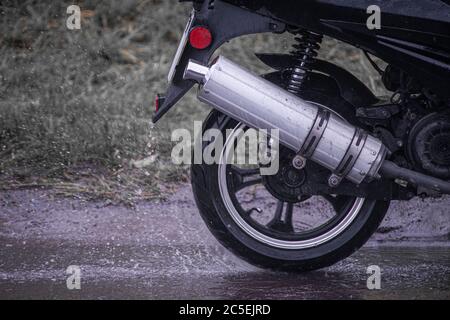 Moto ciclomotore si muove attraverso un puddle su una strada bagnata sotto la pioggia. Lo spruzzo vola dalle ruote. Primo piano Foto Stock