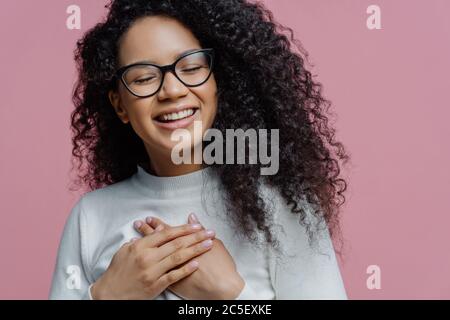 Primo piano di felice sincera donna afro americana mantiene le mani pressate al petto, esprime sentimenti sinceri, fa gesto di gratitudine, si sente grato f Foto Stock