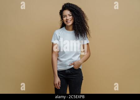 Foto di slim allegra donna afroamericana con capelli ricci, sorride felicemente, essere di buon umore, indossa maglietta bianca e jeans, tiene la mano in tasca Foto Stock