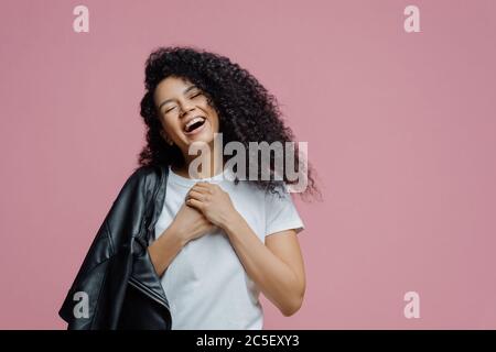 La donna con pelle scura e molto gioiosa tiene insieme le mani e ride sinceramente, chiude gli occhi e si sente molto felice, mostra denti bianchi perfetti, indossa t-shirt e. Foto Stock