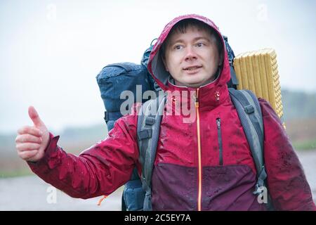 Un ragazzo con uno zaino si trova vicino alla strada, prendendo un giro. Voti turistici per la strada sotto la pioggia Foto Stock