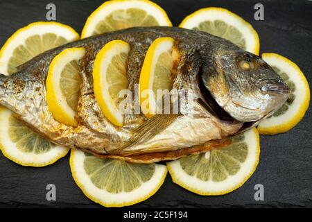 Pesce dorado cotto al forno e limone giacciono su una tavola di ardesia di pietra nera Foto Stock