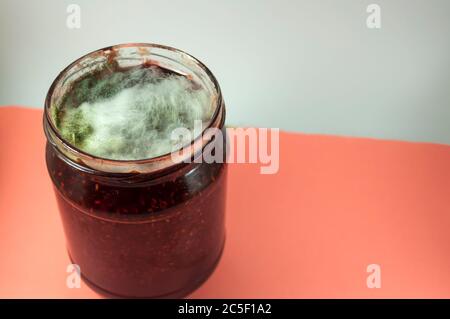 Si può vedere mezzo vaso senza coperchio con confettura di lamponi, che è completamente coperto con fili di stampo bianco e grigio. Foto su sfondo bianco-blu Foto Stock