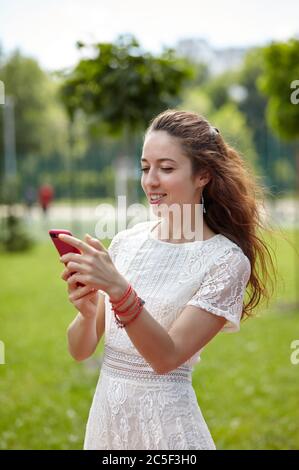 Giovane donna in abito che tiene il telefono cellulare nel parco estivo. La ragazza caucasica gode di uno stile di vita estivo luminoso. Foto Stock