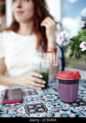 Una giovane donna tiene una tazza di plastica con un mojito in una città estate cafe.tazza di carta di caffè e smartphone sul tavolo Foto Stock