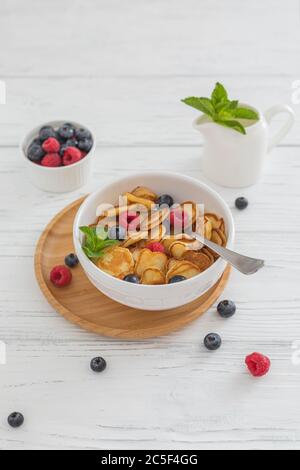 Prima colazione ciotola bianca con minuscoli cereali pancake con mirtilli, lamponi e menta su sfondo di legno Foto Stock