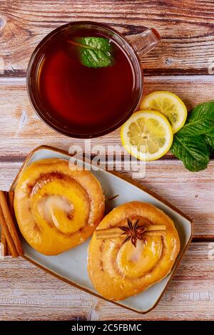 Involtini di cannella fatti in casa con smalto e tazza di tè con limone e menta su sfondo di legno. Foto Stock