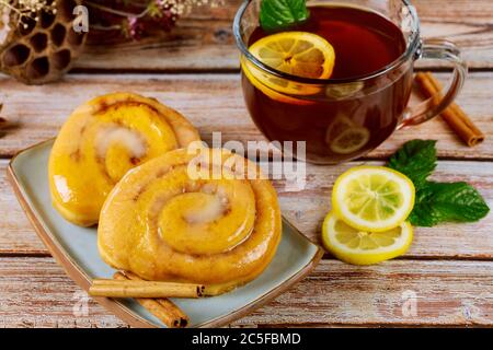 Involtini di cannella con smalto e tazza di tè con limone e menta su sfondo di legno. Foto Stock