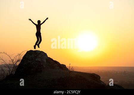 Silhouette di donna escursionista che salta da solo su roccia vuota al tramonto in montagna. Turista femminile che alza le mani in piedi sulla scogliera nella natura serale. Foto Stock